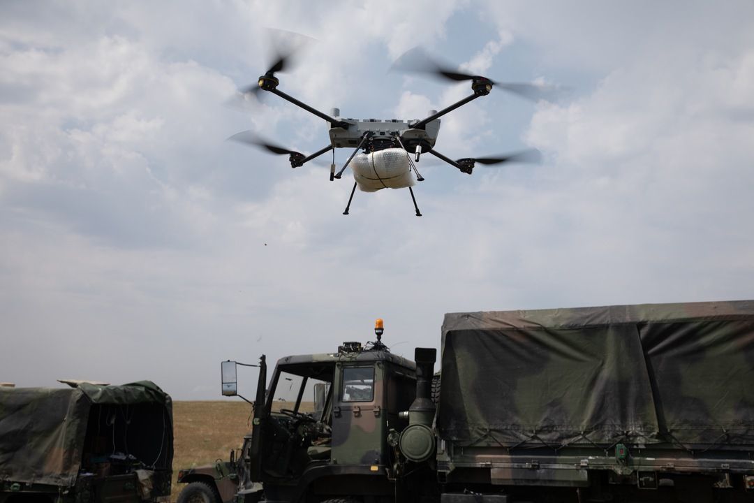 Military multirotor drone carrying a payload hovering above army trucks