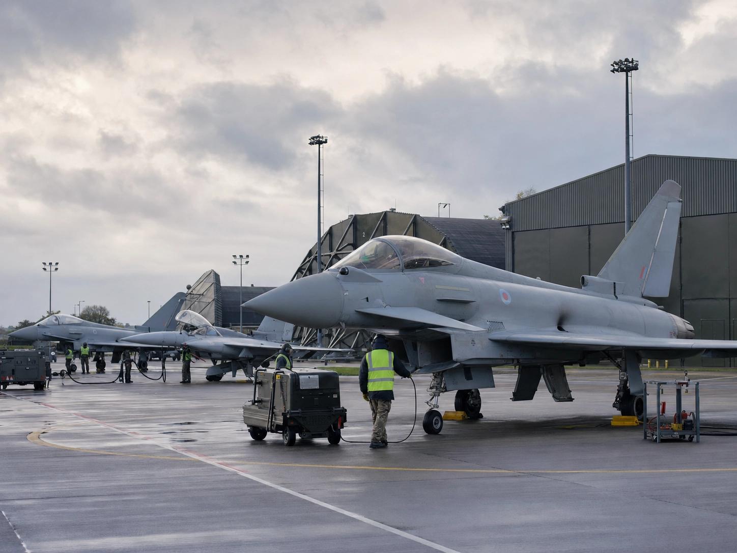 RAF Typhoon fighter jets on a wet flightline as ground crew service them beside a hangar