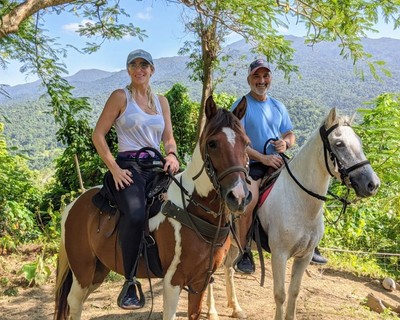 A smiling couple on horseback pose under tropical trees with breathtaking mountain and rainforest views during their La Vista Rides experience in El Yunque, Puerto Rico.