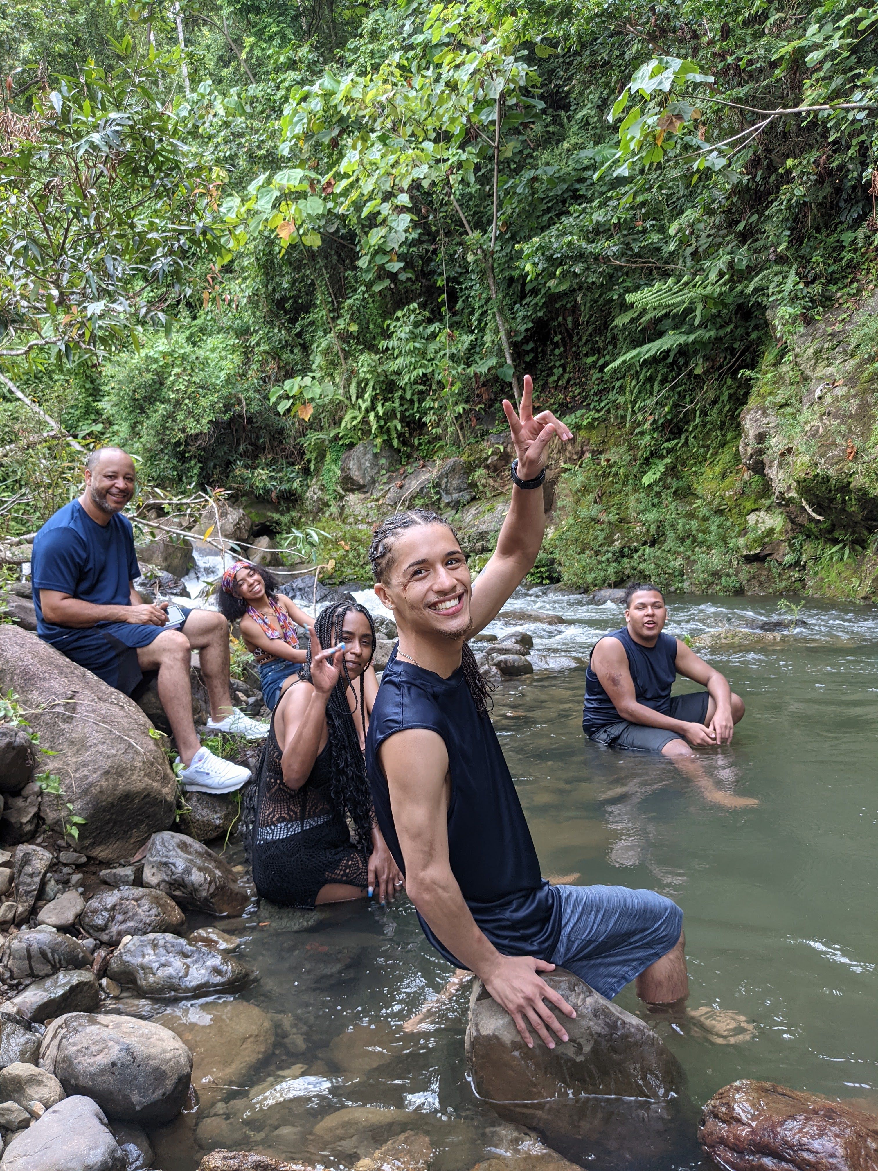 A smiling group of friends relaxing in a shallow rainforest river, surrounded by rocks and lush green jungle.