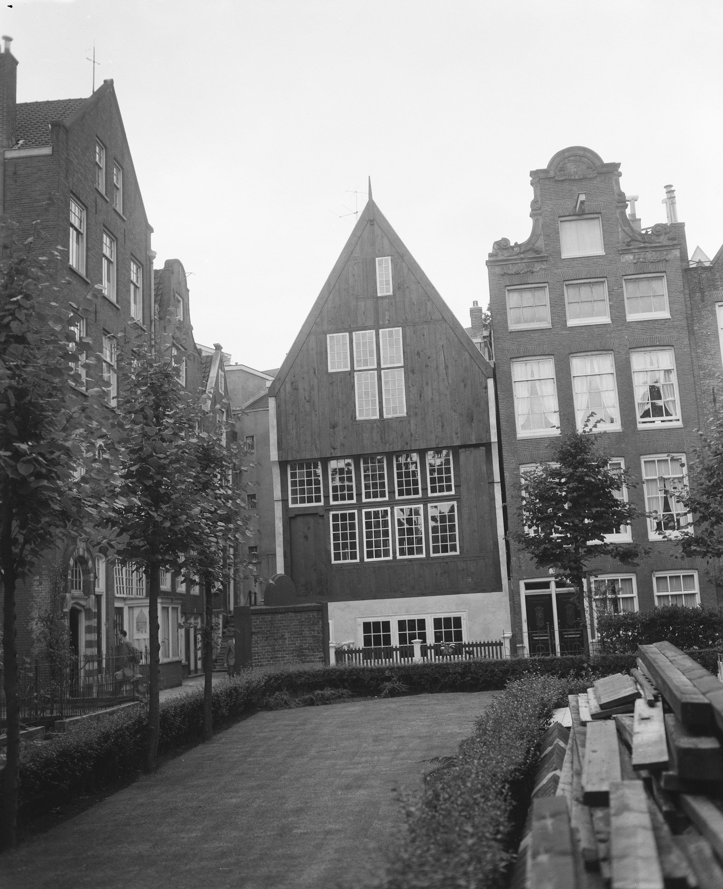 Fotografia em preto e branco de 1957 da fachada de madeira do Houten Huys, no Begijnhof, em Amsterdã, entre casas de tijolo.