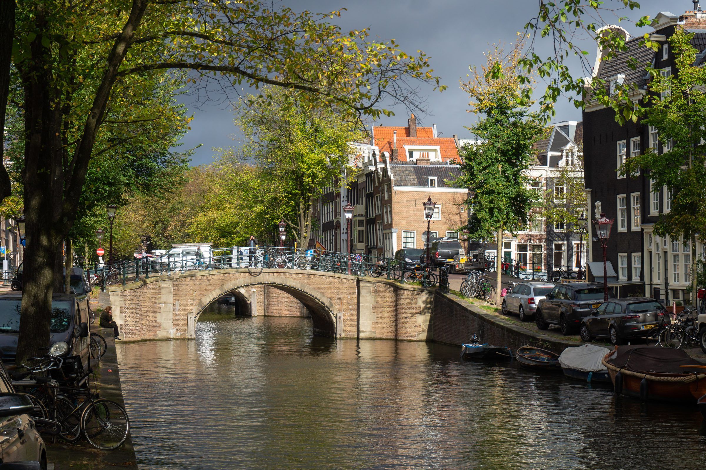O canal Reguliersgracht, em Amsterdã: fachadas de tijolo iluminadas pelo sol sob um céu carregado, uma ponte de pedra em arco sobre a água, barcos atracados e árvores; carros estacionados no cais à direita.
