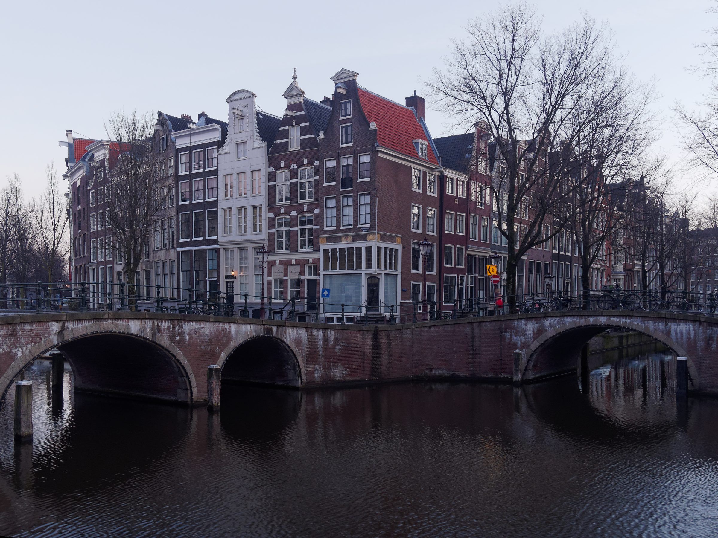 Encontro da Keizersgracht com a Leidsegracht numa manhã de inverno: pontes em arco sobre os canais, árvores sem folhas e fachadas de tijolo ao longo das margens.