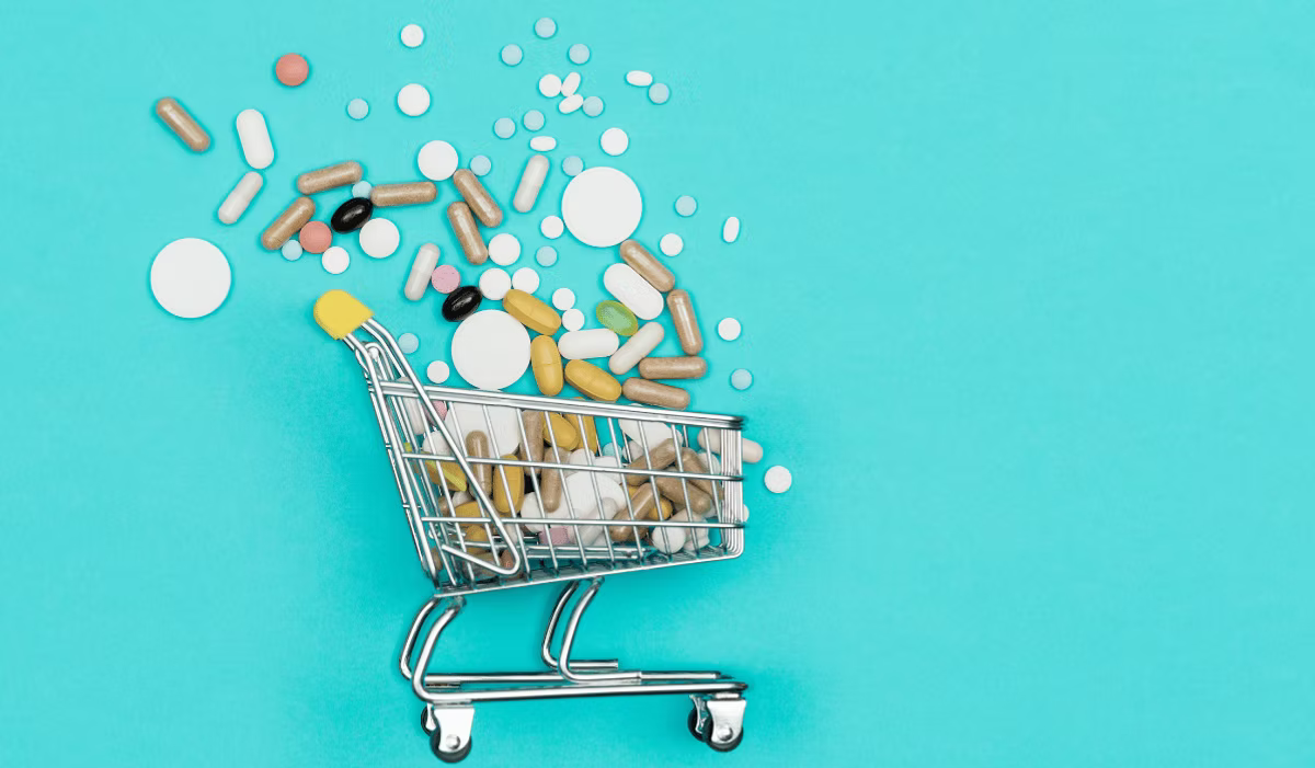 A miniature shopping trolley on a turquoise background, overflowing with assorted pills, capsules and tablets that spill out across the surface.
