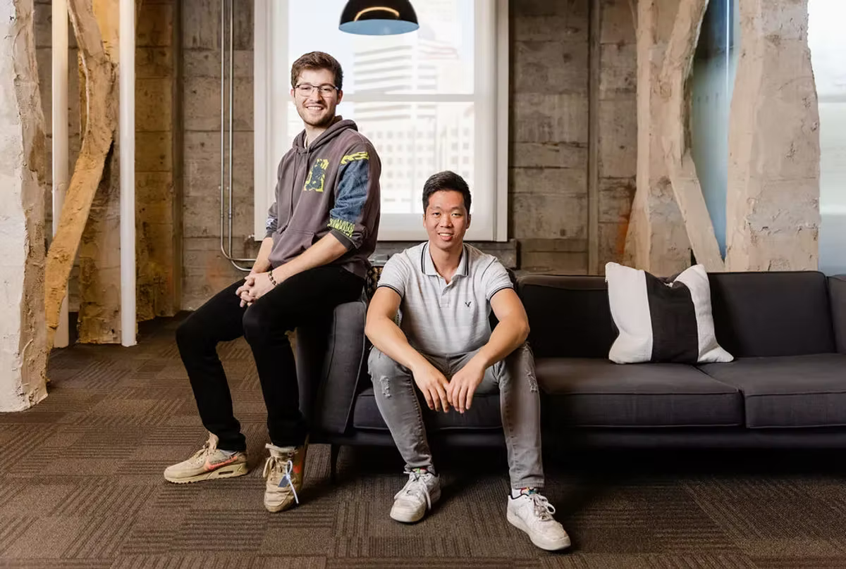 Two young founders posed on and beside a dark sofa in a modern office with concrete pillars and a city view.