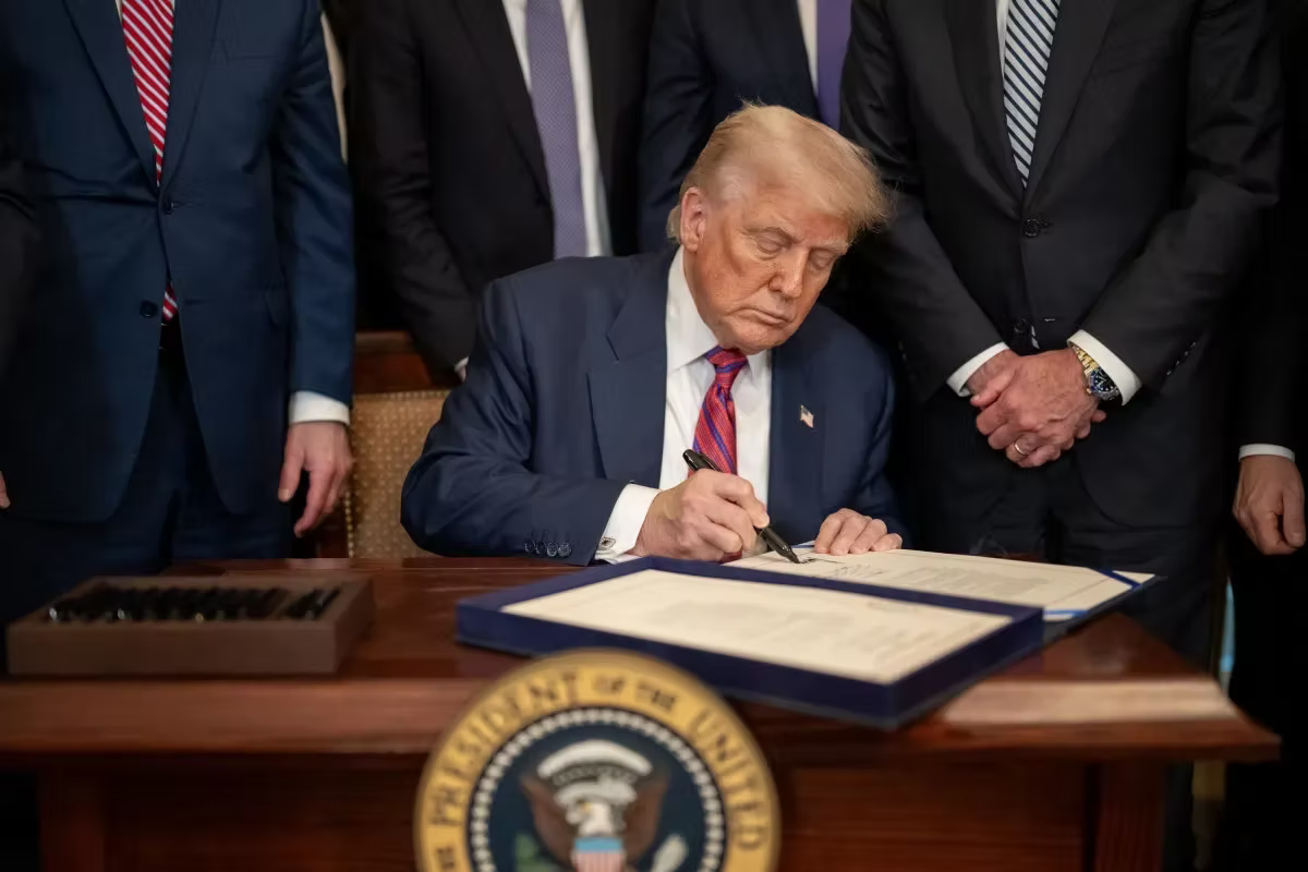 Donald Trump seated at a desk signing a document, flanked by men in suits, with the presidential seal on the desk front.