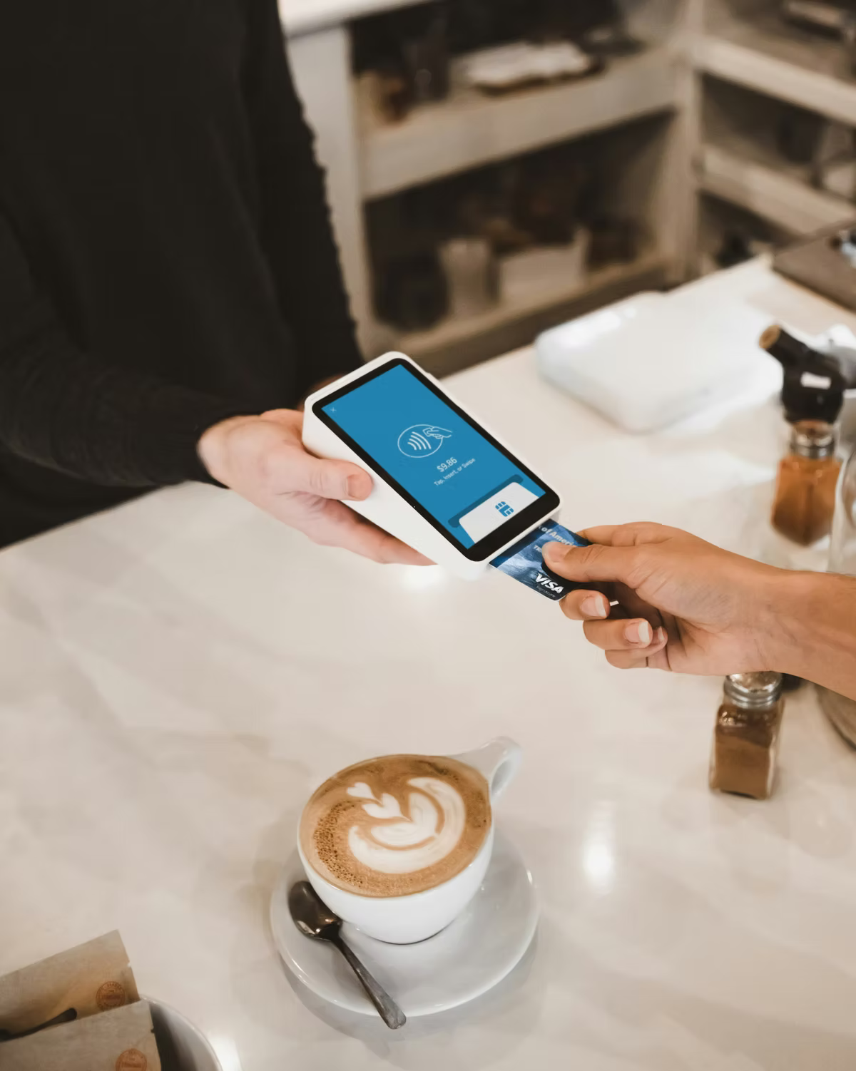 A customer taps a Visa card against a handheld card reader held out by a cafe worker, a latte in a white cup on the counter below.