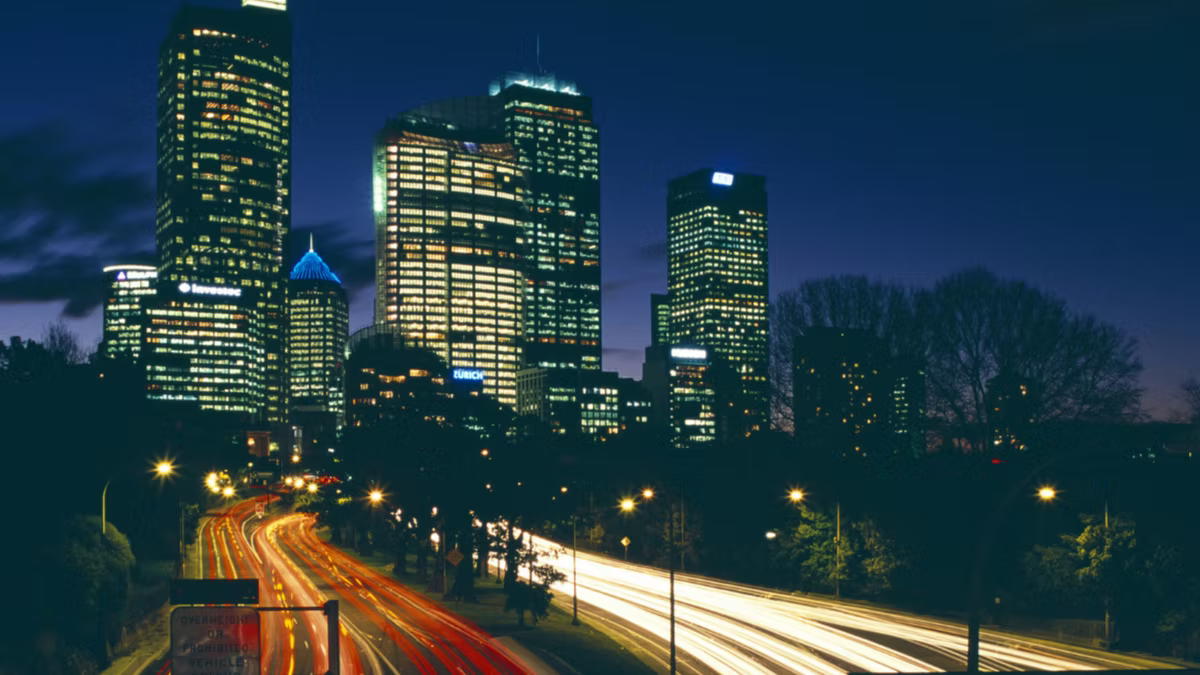 A long-exposure night view of a city, office towers lit behind and light trails from traffic streaking along the road below.