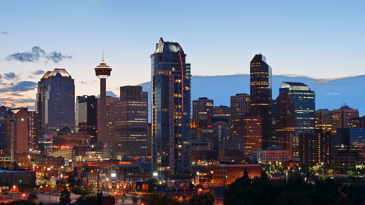The Calgary skyline at dusk, the Calgary Tower among lit office towers under a pale blue and orange sky.