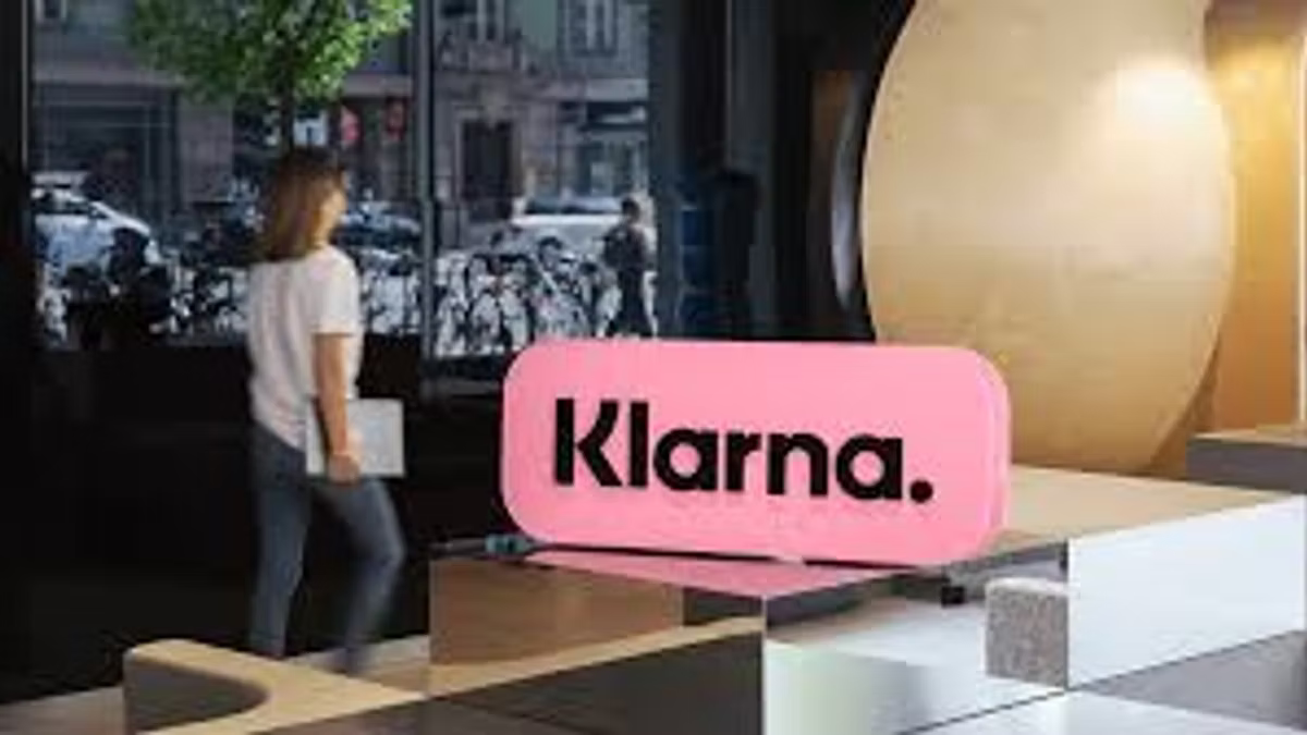 A large pink Klarna sign on a plinth inside a bright office lobby, with a woman walking past the window behind it.