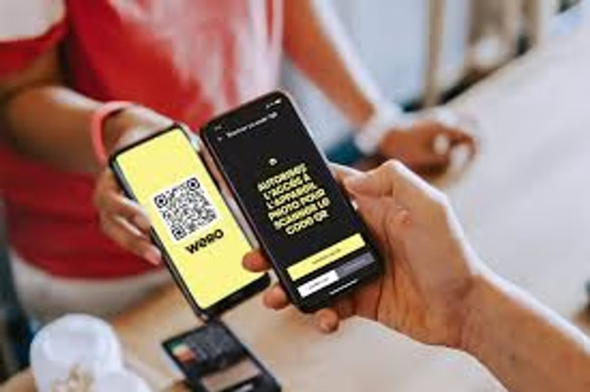 Two people hold phones together to make a Wero payment: one screen shows a yellow Wero QR code, the other asks in French for permission to use the camera to scan it.