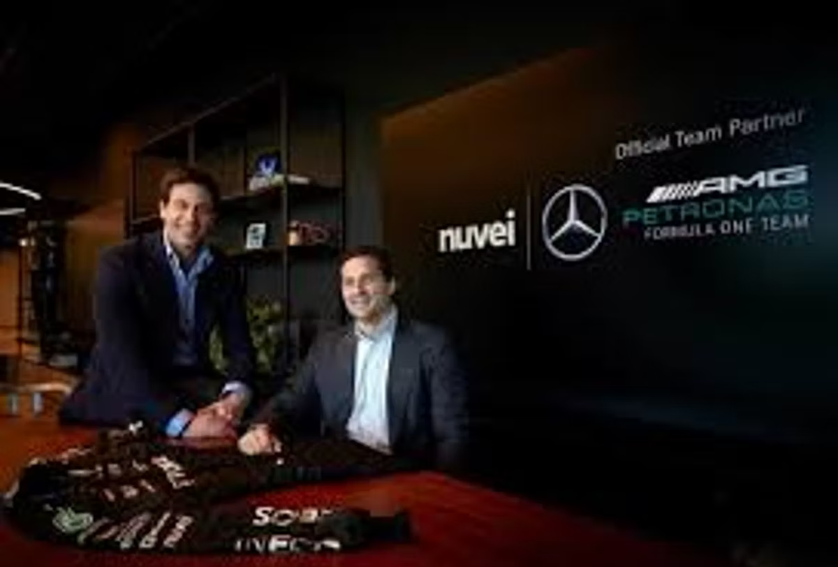 Two men seated at a table with a signed team jacket in front of them, beside a screen reading "Official Team Partner" with the Nuvei and Mercedes-AMG Petronas Formula One Team logos.