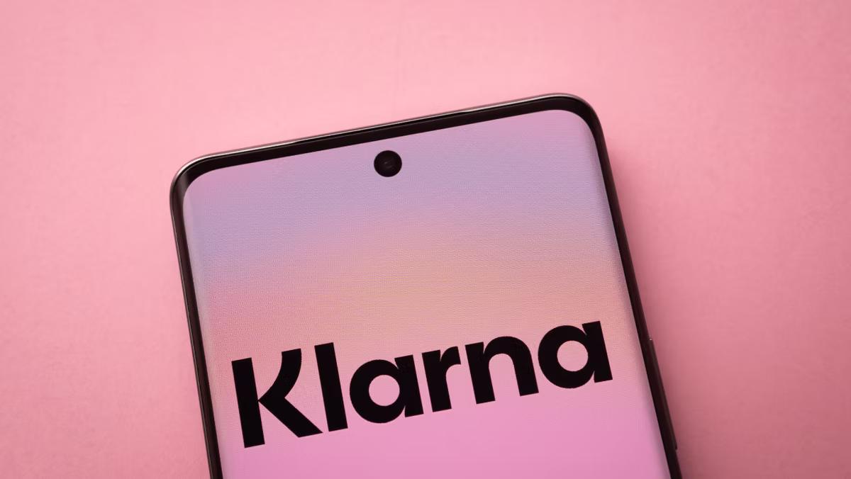 Smartphone in close-up on pink background, screen displays "Klarna." Soft pastel colors evoke a calm, modern feel.