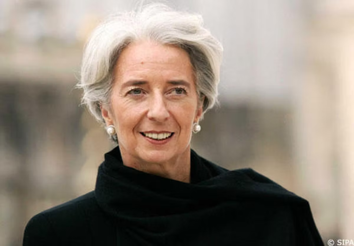 Portrait of a woman with short white hair, wearing a black coat and scarf and pearl earrings, smiling outdoors.