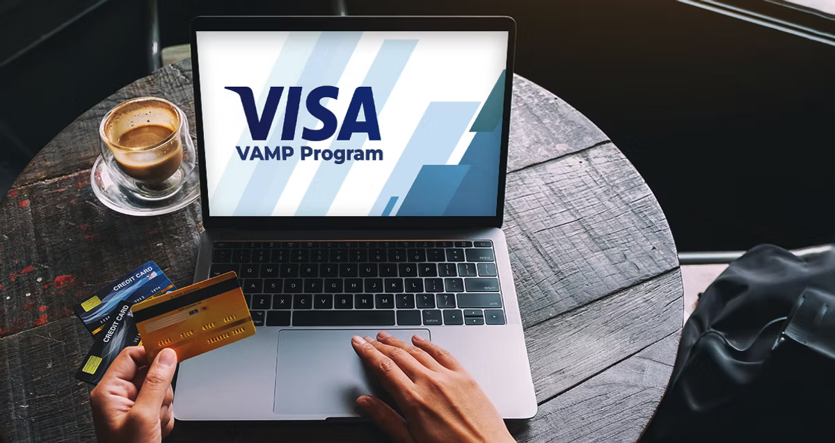 Hands hold a credit card near a laptop displaying "VISA VAMP Program." A cup of coffee and more cards sit on the rustic wooden table.