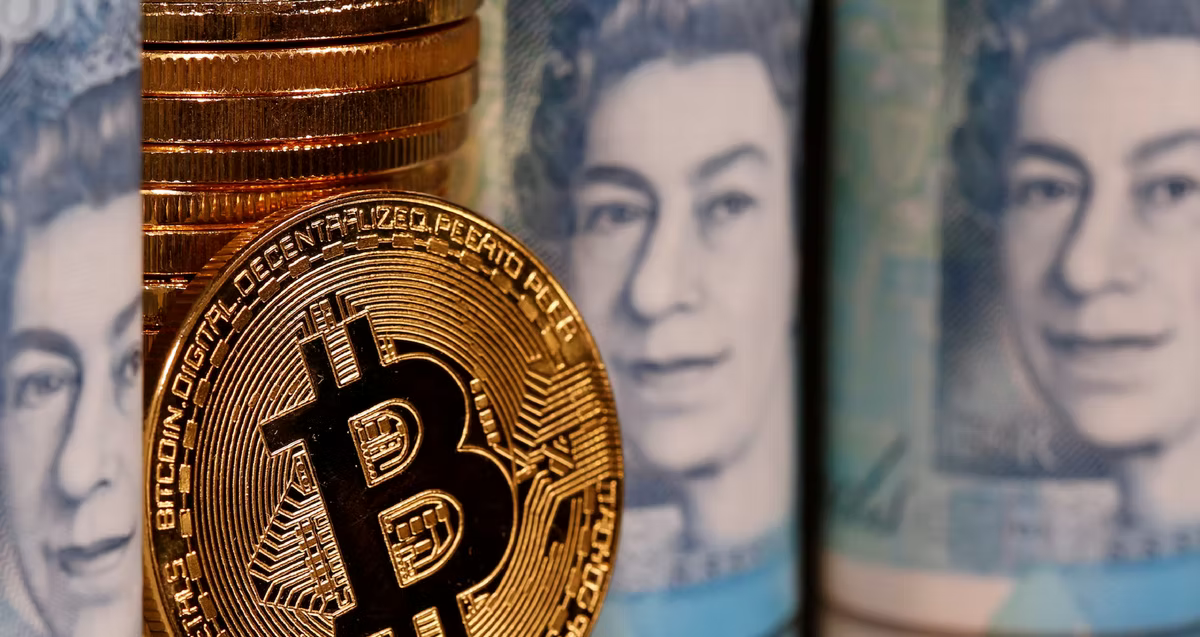 A gold Bitcoin coin propped against a stack of coins in front of rolled British banknotes.