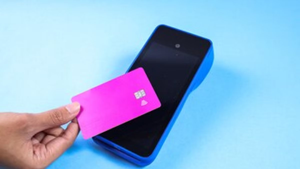 A hand holds a plain pink contactless payment card up to a blue card terminal lying on a pale blue background.