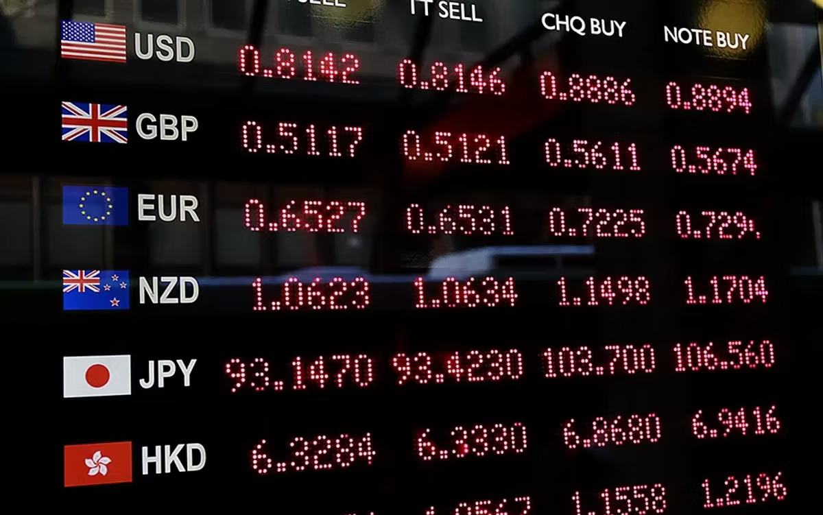 A currency exchange board glowing in red, listing sell and buy rates against flags for USD, GBP, EUR, NZD, JPY and HKD.