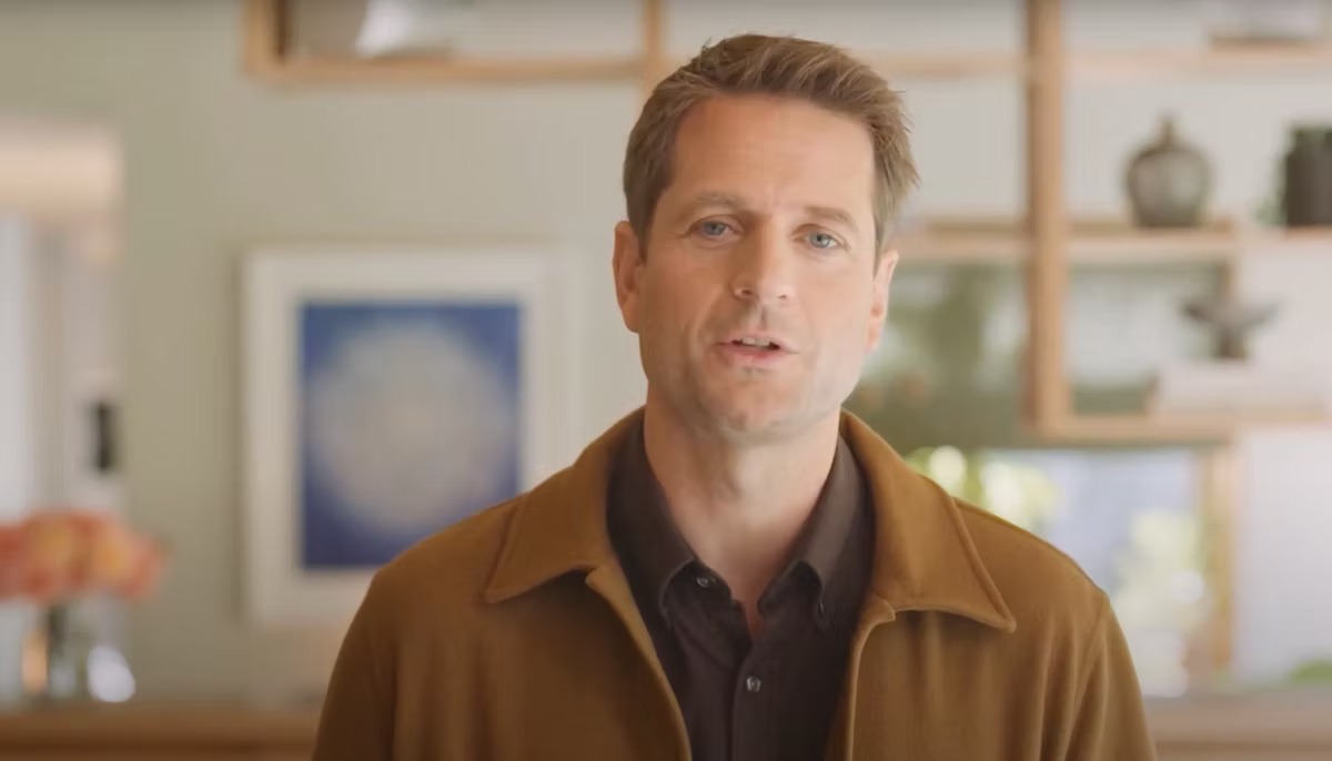 Klarna CEO Sebastian Siemiatkowski in a brown jacket speaks in a room with shelves and abstract art in the background. Soft lighting, neutral tones.