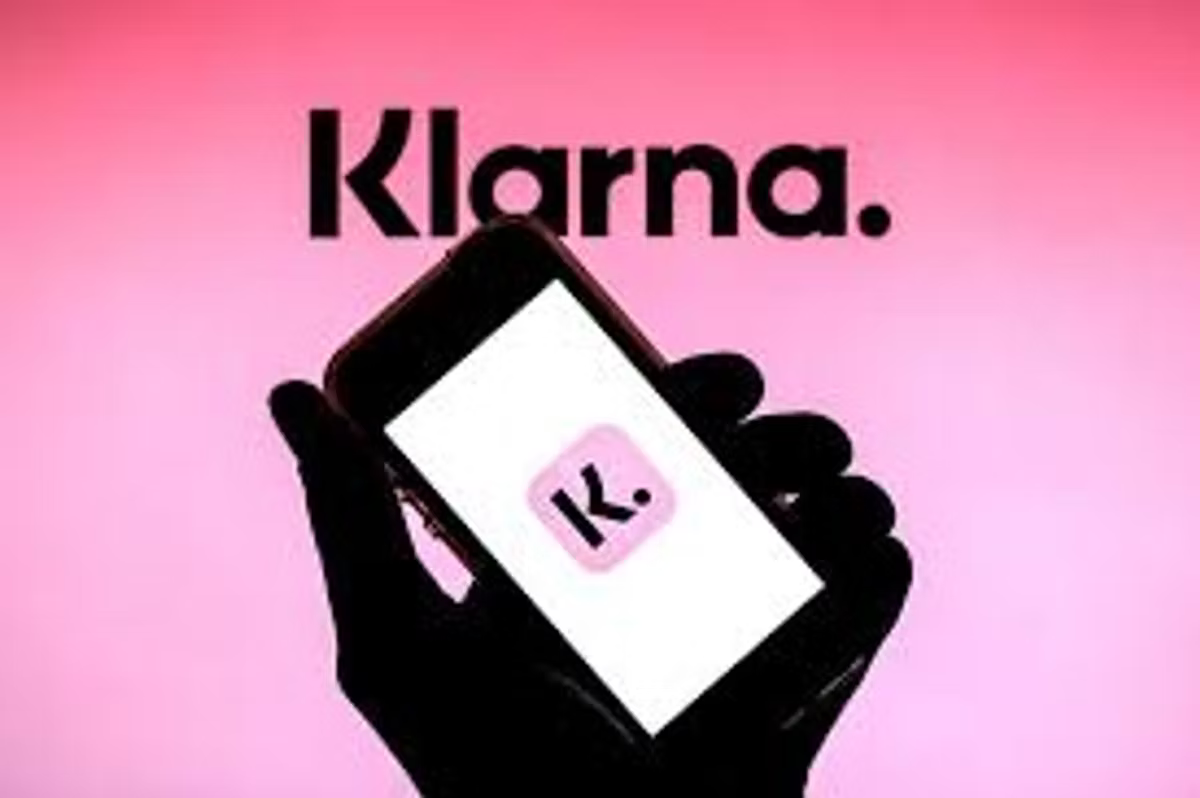 A silhouetted hand holding a phone showing the Klarna app icon, against a pink background with the Klarna wordmark above.