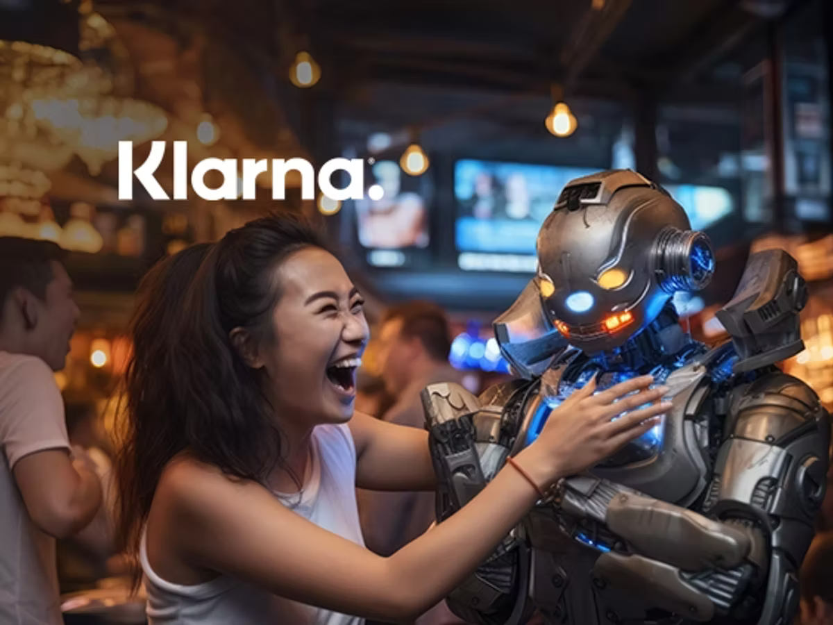 The Klarna wordmark over a photograph of a laughing woman in a bar playfully grappling with a humanoid robot lit with blue and orange lights.