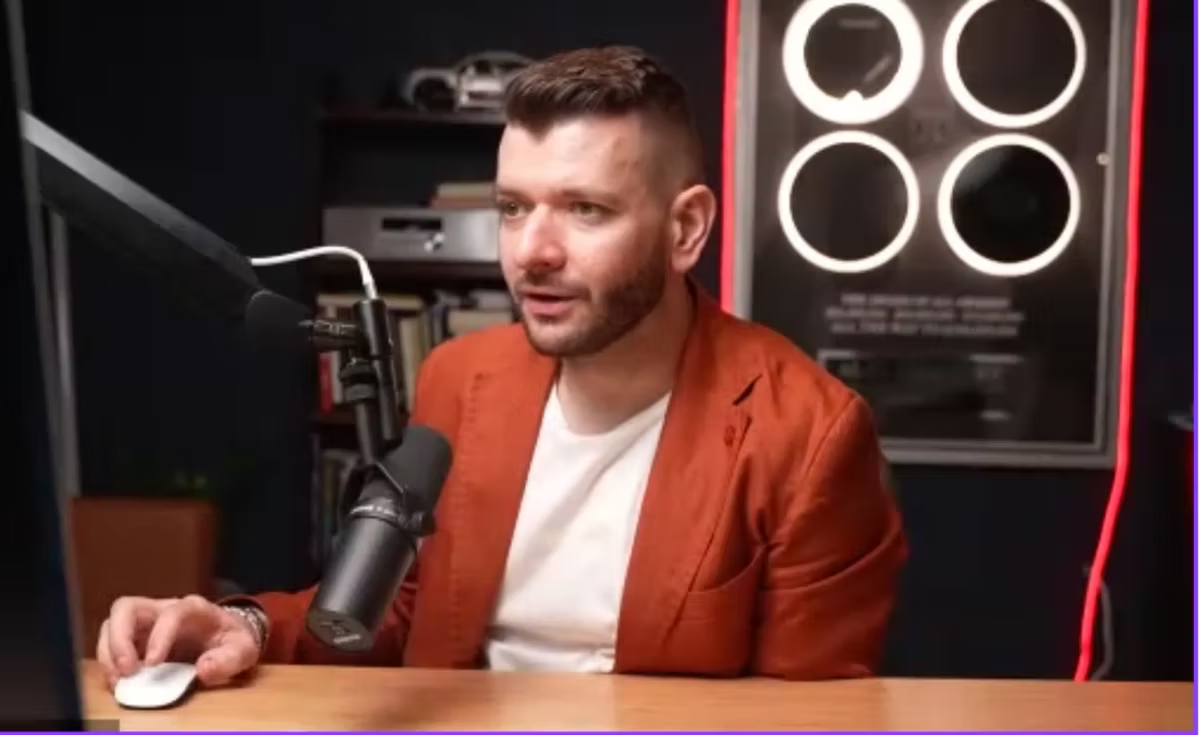 A man in a rust-coloured blazer speaking into a studio microphone at a desk, with shelving and ring lights behind him.
