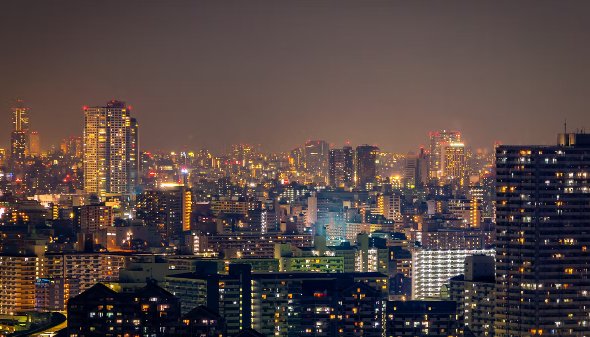 A dense city skyline at night, tower blocks and offices lit orange and white under a hazy sky.