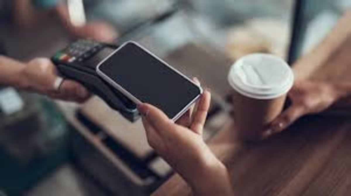 A customer holds a smartphone up to a card terminal held by a member of staff, a takeaway coffee cup on the counter beside them.
