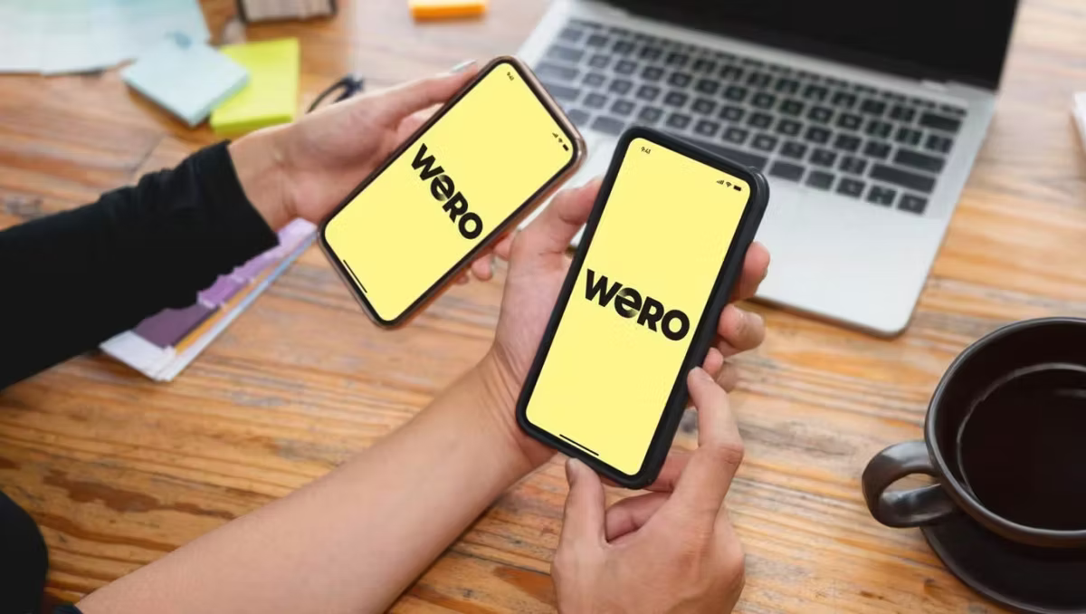 two people holding phones with the word WERO on the center of their screens. there is a wooden table in the background with a laptop and coffee on it.