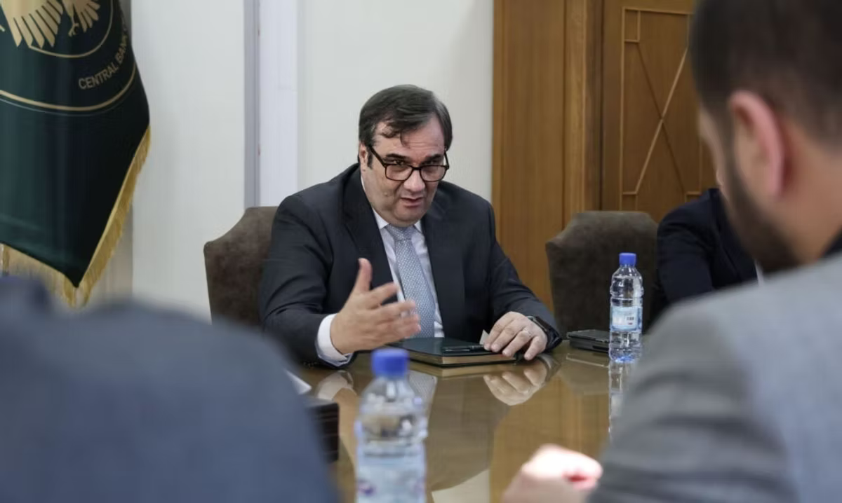 Syrian Central Bank Governor AdbulKader Husrieh in suit gestures during a meeting at a table with water bottles. Background features a flag and wood paneling, conveying a formal mood.