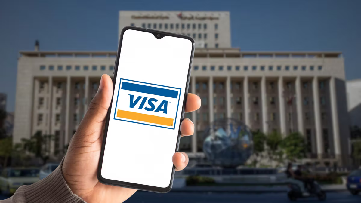 Hand holding phone with Visa logo on screen, in front of a large cream-colored building with columns. Bright, sunny day.