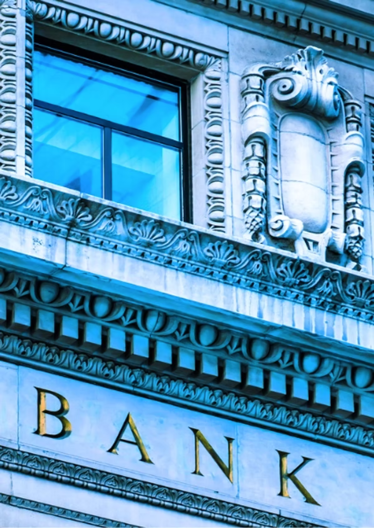 Looking up at the ornate stone facade of a classical bank building, the word BANK in gold lettering across it, tinted blue.