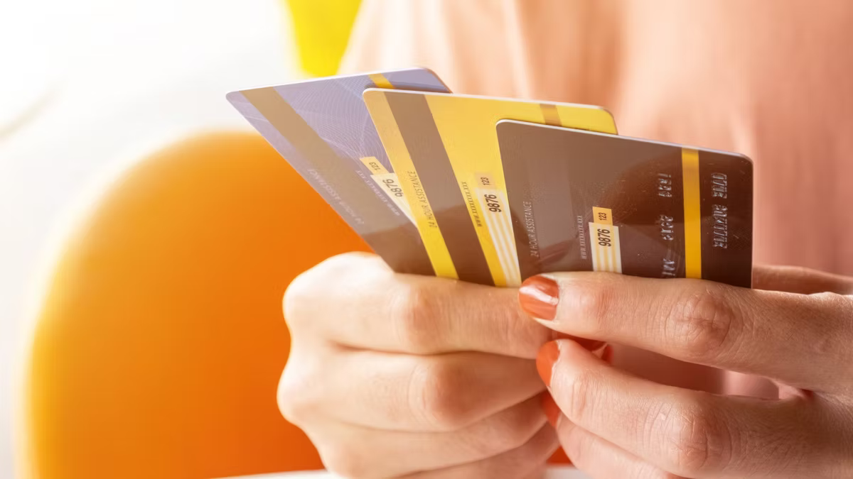 A person holds a fan of three payment cards in purple, yellow and grey, seen close up against a soft orange background.