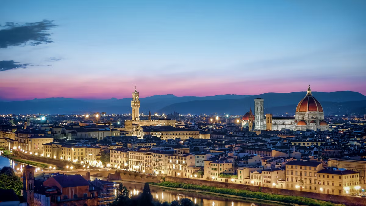 Florence at dusk, the Duomo and Palazzo Vecchio tower lit above the rooftops with the Arno in the foreground under a pink sky.