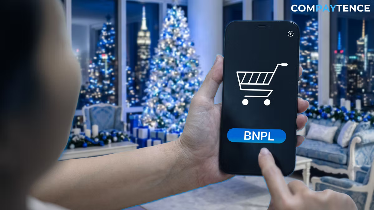 Compaytence hero card showing a hand holding a phone displaying a shopping-cart icon and a blue BNPL button, in a room with lit Christmas trees and a city view behind.