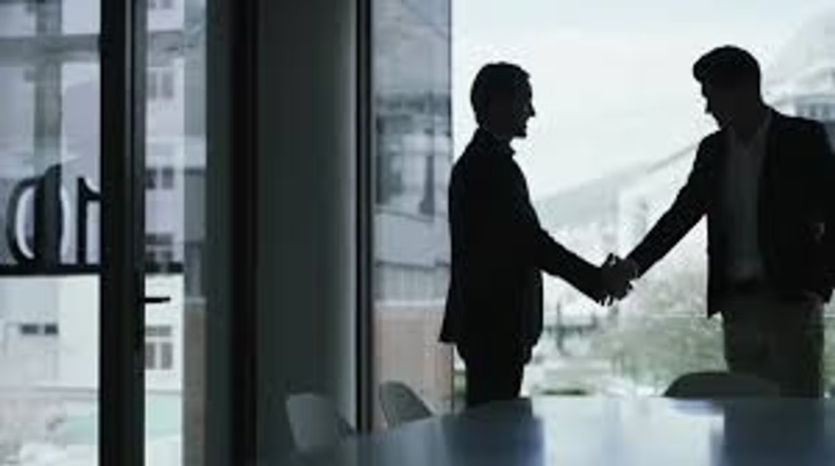 Two people in suits shake hands, silhouetted against an office window with a city view behind them.