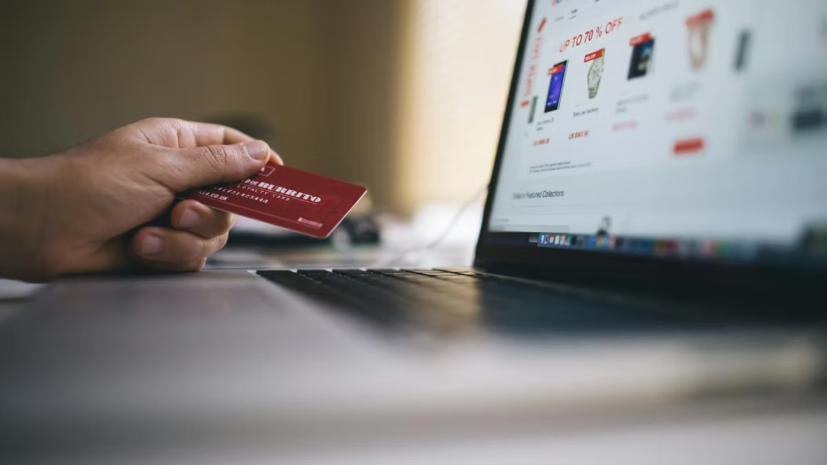 A hand holds a red credit card in front of an open laptop showing an online store banner advertising up to 70% off.