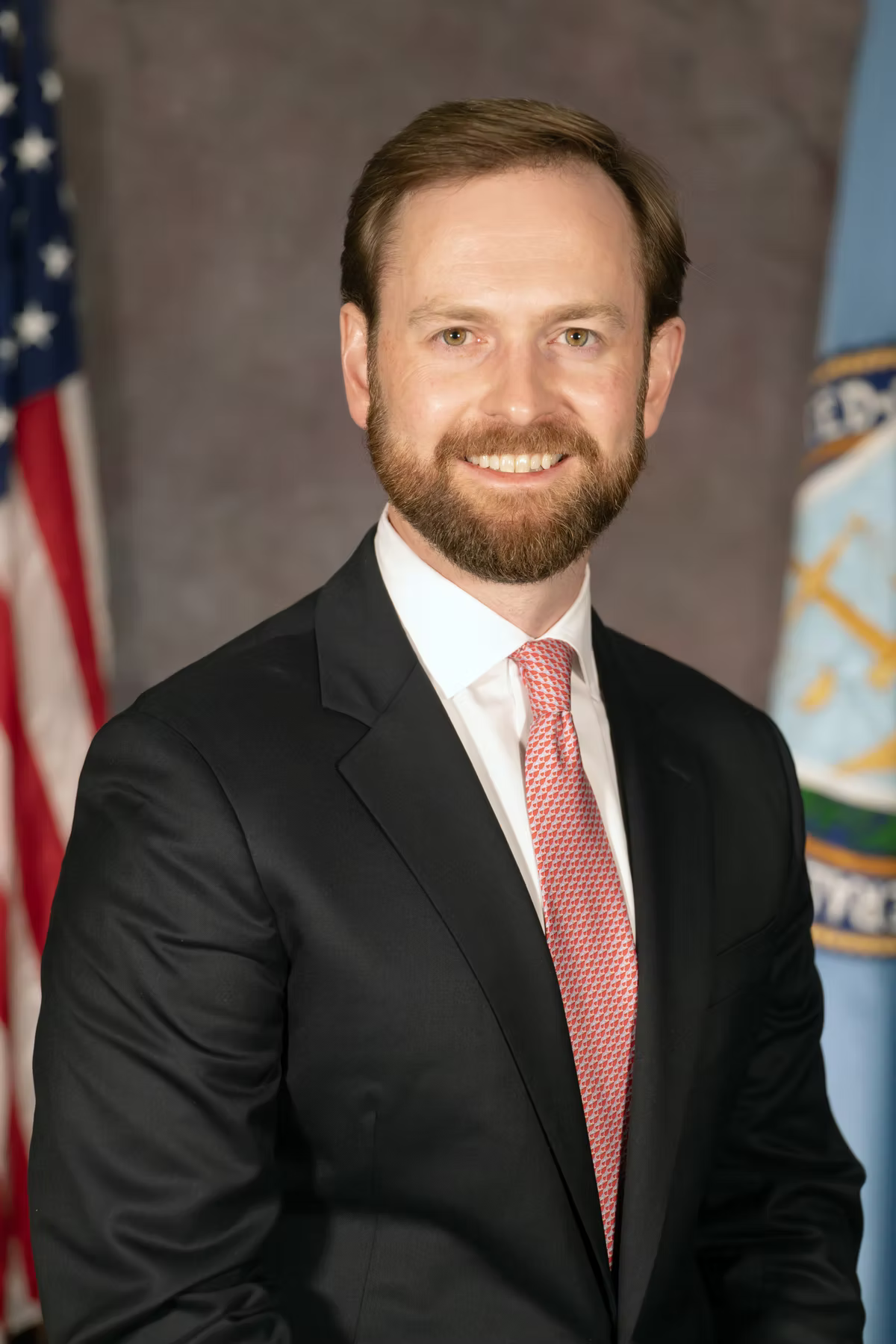 Official portrait of a bearded man in a dark suit and red patterned tie, photographed between a United States flag and a federal agency flag.