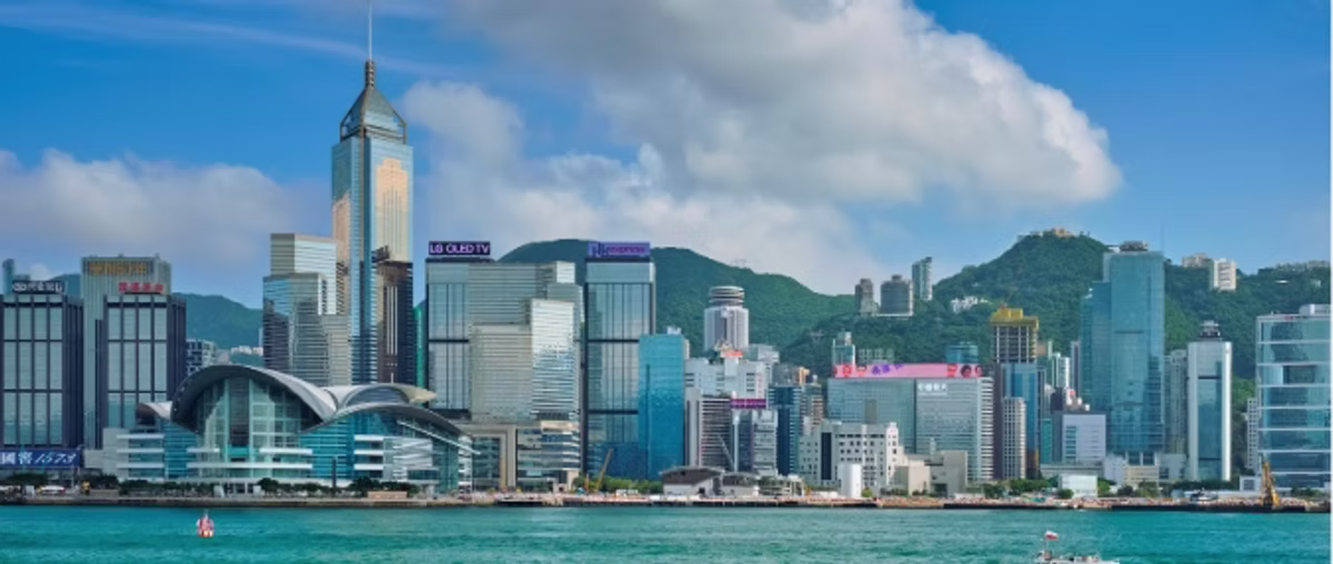 The Hong Kong waterfront skyline seen across Victoria Harbour, towers rising in front of green hills under a blue sky.