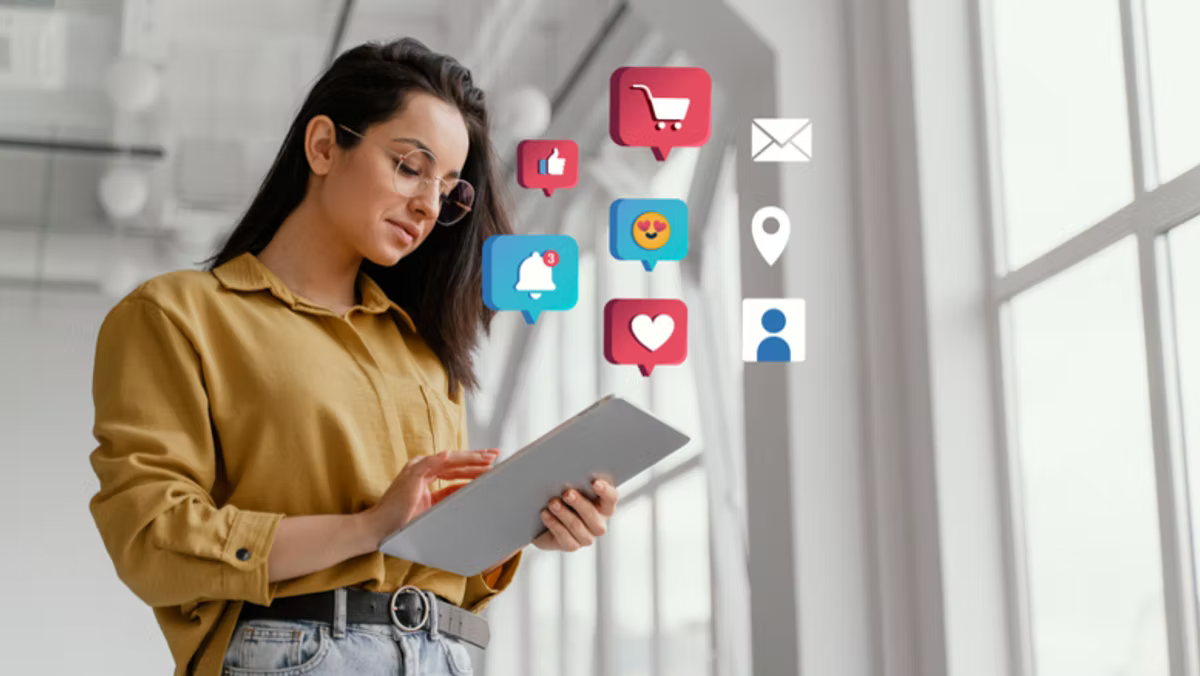 A woman in a mustard shirt uses a tablet by a window, surrounded by floating social media icons: a cart, thumbs up, notification bell, heart, smiling face, envelope, location pin and profile.