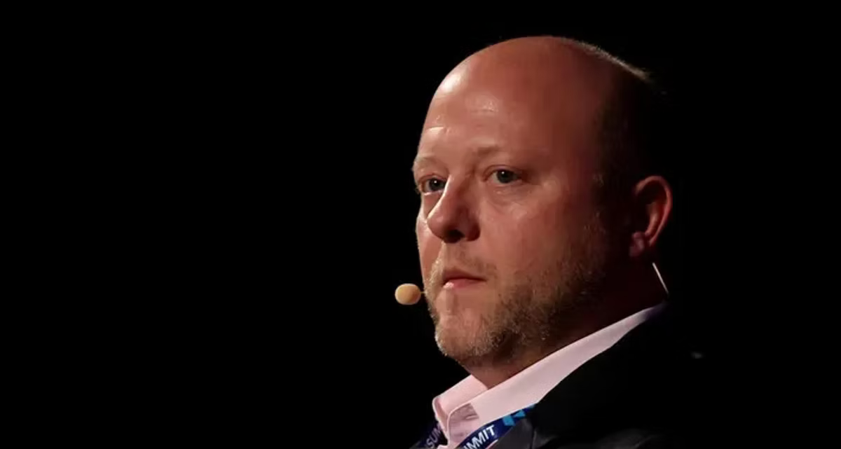A bald man in a dark jacket wearing a stage microphone, photographed in profile against a black conference backdrop.