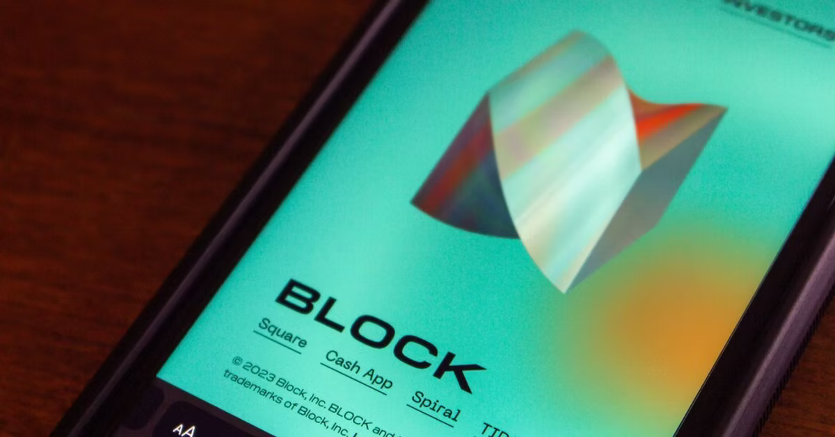 Close-up of a smartphone screen displaying a teal 3D shape and the text "BLOCK" with related companies. Set on a wooden surface.