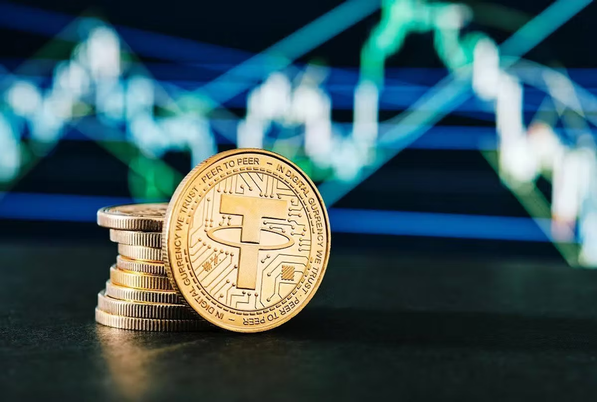 A gold Tether coin standing against a stack of coins, with a blurred green and blue candlestick chart behind.
