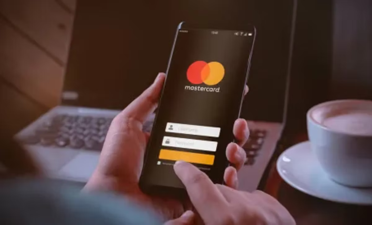 Hands holding a smartphone displaying the Mastercard login screen. A laptop and a cup on a table in the blurred background. Warm lighting.