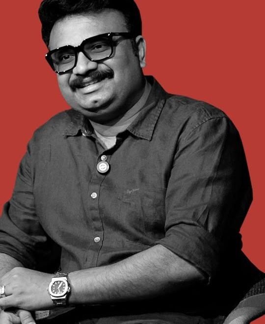 Jis Joy in glasses and a dark shirt, seated against a red backdrop