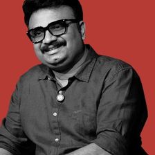 Jis Joy in glasses and a dark shirt, seated against a red backdrop