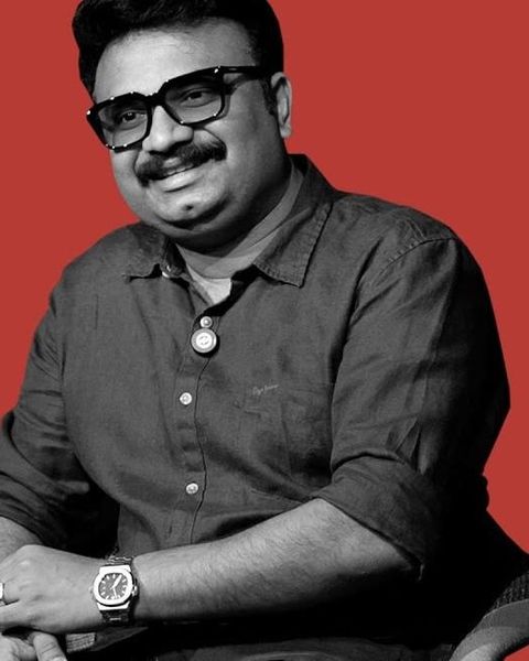 Jis Joy in glasses and a dark shirt, seated against a red backdrop