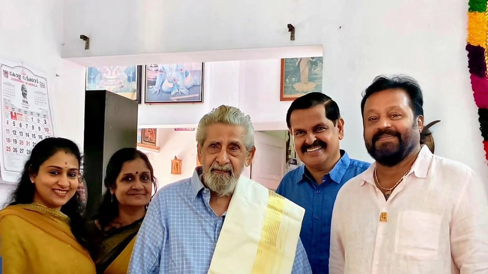 Veteran actor Madhu, in a blue shirt and gold-bordered shawl, stands at home with Suresh Gopi and family members