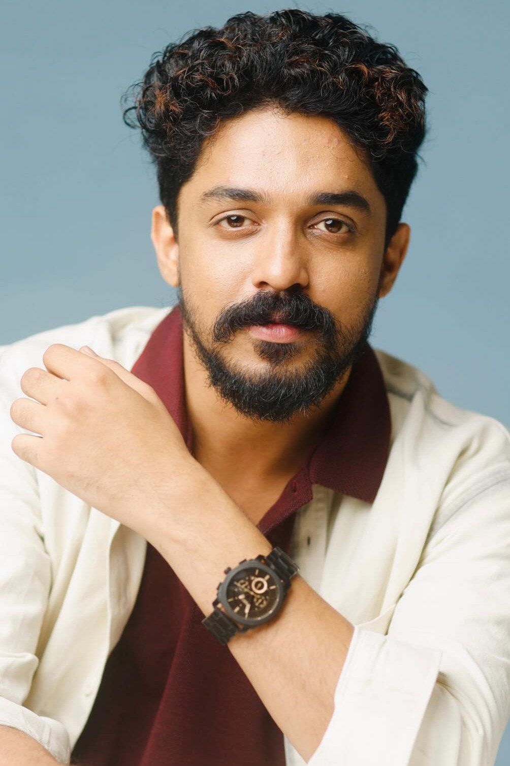 Sagar Surya with curly hair and a beard in a cream shirt over a maroon top, blue backdrop portrait