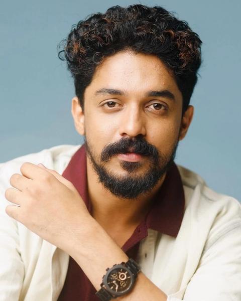 Sagar Surya with curly hair and a beard in a cream shirt over a maroon top, blue backdrop portrait